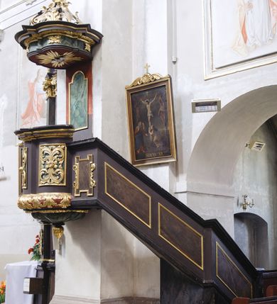 Zdjęcie nr 1: Ambona wisząca, przyścienna, umieszczona na ścianie łuku tęczowego, z parapetem dziś nieistniejących schodów od strony prezbiterium i usytuowanej przy nim kaplicy. Kosz ambony założony na rzucie trójliścia z trójkątnymi występami, wsparty na puklowanej konsoli z podwieszoną szyszką. Dolna część ambony wybrzuszona, pokryta rozłożystymi liśćmi akantu, kosz prosty na rzucie odcinków koła z prostokątnymi płycinami wypełnionymi liśćmi akantu, zakończony profilowanym gzymsem. Zaplecek z profilowaną płyciną zakończoną łukiem półkolistym nadwieszonym. Nad koszem baldachim powielający rzut kosza, zwieńczony silnie wystającym gzymsem, z podwieszoną w podniebieniu gołębicą Ducha Świętego na tle glorii promienistej. Nad baldachimem kabłąki w formie spływów wolutowych opatrzonych liśćmi akantu, podtrzymujące koronę zamkniętą z kulą i krzyżem. Parapet schodów z dekoracją w postaci trzech płycin w kształcie równoległoboku obwiedzionych profilowanymi ramami. Płycina w polu łączącym parapet schodów z podestem przed amboną w formie stojącego prostokąta z uszakami, ujętego motywami roślinnymi. Ambona w kolorze czarnym, z zielonymi i czerwonymi imitacjami marmoryzacji, ze złoconymi ornamentami. 