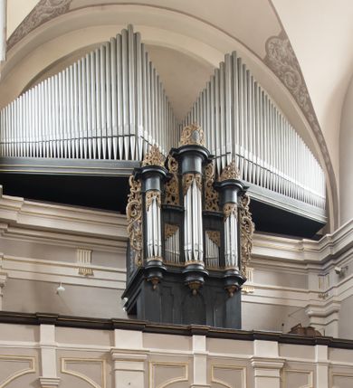 Zdjęcie nr 1: Część zachodnią nawy kościoła zamyka murowany, trójarkadowy chór muzyczny. Sklepiony jest on krzyżowo, dostępny od południa, wsparty na dwóch filarach opiętych pilastrami od strony wschodniej, oraz dwóch półfilarach przylegających do ścian. Parapet chóru zakończony jest uproszczonym belkowaniem dźwiganym przez półfilary oraz wąskimi pilastrami ponad arkadami, umieszczonymi na ich osi. Belkowanie zostało wyłamane ponad filarami i pilastrami. Powyżej arkad, w przestrzeniach między pilastrami umieszczone są płyciny dopasowane kształtem do pola, które zajmują. Otoczone są profilowaną, złoconą ramą.
Prospekt organowy wykonany z drewna, wsparty na wysokim cokole, z trzema wieżyczkami na rzucie półkolistym, osadzone na postumentach z podwieszonymi rozetami i szyszkami. Między wieżyczkami, z których środkowa jest wyższa od bocznych. Zarówno wieżyczki jak i ścianki z piszczałkami są zwieńczone odcinkami gzymsów. Cały instrument zdobiony ornamentem małżowinowo-chrząstkowym, występującym w formie zwieńczeń, uszu i dekoracji górnej części grup piszczałek. Elementy ornamentalne złocone. Korpus prospektu zdobią trzy płyciny. Środkowa, dwa razy szersza płycina ma kształt stojącego prostokąta zakończonego łukiem nadwieszonym. Z kolei dwie symetryczne płyciny skrajne zakończone są łukiem dwudzielnym. Wszystkie obejmuje profilowana rama.