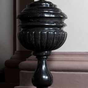 Zdjęcie nr 1: Chrzcielnica usytuowana na postumencie na rzucie półkolistym i schodkowej, kwadratowej płycie. Jej trzon w typie gruszkowym dźwiga puklowaną, półkulistą czarę z profilowanym, ćwierćkolistym w przekroju wylewem. Pokrywa drewniana, bogato profilowana, malowana na czarno, zwieńczona złoconą kulą z krzyżem. 

