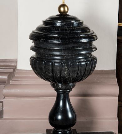 Zdjęcie nr 2: Chrzcielnica usytuowana na postumencie na rzucie półkolistym i schodkowej, kwadratowej płycie. Jej trzon w typie gruszkowym dźwiga puklowaną, półkulistą czarę z profilowanym, ćwierćkolistym w przekroju wylewem. Pokrywa drewniana, bogato profilowana, malowana na czarno, zwieńczona złoconą kulą z krzyżem. 

