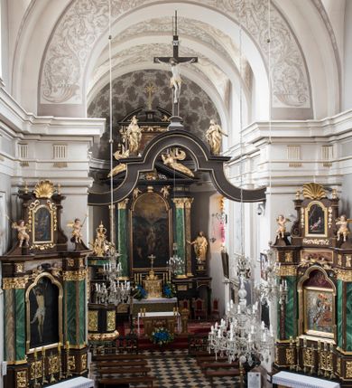 Zdjęcie nr 6: Na belce tęczowej o zarysie łuku wklęsło-wypukłego z uskokiem usytuowano krzyż z figurą Chrystusa i figury Marii i św. Jana Ewangelisty. Chrystus został przedstawiony frontalnie, w lekkim zwisie, z głową na wysokości dłoni, z nogami lekko ugiętymi w kolanach, skrzyżowanymi stopami, prawa na lewej, przybitymi do krzyża jednym gwoździem. Ręce rozłożone szeroko na ramionach krzyża, dłonie z lekko zgiętymi palcami są przebite gwoździami. Sylwetka Chrystusa jest delikatnie wygięta, głowa bezwładnie opada na prawe ramię. Jego twarz jest pociągła, nos prosty, wąski, oczy małe, zamknięte. Twarz okala szpiczasta, brązowa, krótka broda. Głowę przykrywają gęste, ciemne włosy o falistych, długich puklach zakrywające ucho i opadające na szyję oraz złota korona cierniowa. Ciało opracowane poprawnie pod względem anatomii, ale modelowane delikatnie, z lekko zarysowaną muskulaturą. Na biodrach krótkie perizonium, ze swobodnie opadającym wzdłuż prawego uda końcem materiału. W prawym boku Chrystusa widoczna jest rana od włóczni, z której wypływa krew, podobnie, jak z ran powstałych przez koronę cierniową, czy w miejscach przebitych gwoździami. Belka łuku tęczowego w kolorze brązowym, z fragmentami profilowania złoconymi. Rzeźba Chrystusa polichromowana naturalistycznie, ze złoconym perizonium oraz koroną cierniową i czerwonymi strużkami krwi.