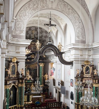 Zdjęcie nr 2: Na belce tęczowej o zarysie łuku wklęsło-wypukłego z uskokiem usytuowano krzyż z figurą Chrystusa i figury Marii i św. Jana Ewangelisty. Chrystus został przedstawiony frontalnie, w lekkim zwisie, z głową na wysokości dłoni, z nogami lekko ugiętymi w kolanach, skrzyżowanymi stopami, prawa na lewej, przybitymi do krzyża jednym gwoździem. Ręce rozłożone szeroko na ramionach krzyża, dłonie z lekko zgiętymi palcami są przebite gwoździami. Sylwetka Chrystusa jest delikatnie wygięta, głowa bezwładnie opada na prawe ramię. Jego twarz jest pociągła, nos prosty, wąski, oczy małe, zamknięte. Twarz okala szpiczasta, brązowa, krótka broda. Głowę przykrywają gęste, ciemne włosy o falistych, długich puklach zakrywające ucho i opadające na szyję oraz złota korona cierniowa. Ciało opracowane poprawnie pod względem anatomii, ale modelowane delikatnie, z lekko zarysowaną muskulaturą. Na biodrach krótkie perizonium, ze swobodnie opadającym wzdłuż prawego uda końcem materiału. W prawym boku Chrystusa widoczna jest rana od włóczni, z której wypływa krew, podobnie, jak z ran powstałych przez koronę cierniową, czy w miejscach przebitych gwoździami. Belka łuku tęczowego w kolorze brązowym, z fragmentami profilowania złoconymi. Rzeźba Chrystusa polichromowana naturalistycznie, ze złoconym perizonium oraz koroną cierniową i czerwonymi strużkami krwi.