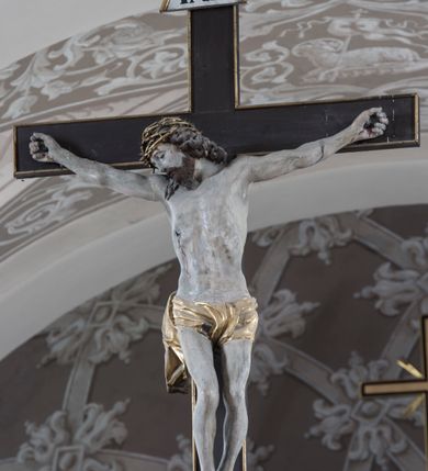 Zdjęcie nr 5: Na belce tęczowej o zarysie łuku wklęsło-wypukłego z uskokiem usytuowano krzyż z figurą Chrystusa i figury Marii i św. Jana Ewangelisty. Chrystus został przedstawiony frontalnie, w lekkim zwisie, z głową na wysokości dłoni, z nogami lekko ugiętymi w kolanach, skrzyżowanymi stopami, prawa na lewej, przybitymi do krzyża jednym gwoździem. Ręce rozłożone szeroko na ramionach krzyża, dłonie z lekko zgiętymi palcami są przebite gwoździami. Sylwetka Chrystusa jest delikatnie wygięta, głowa bezwładnie opada na prawe ramię. Jego twarz jest pociągła, nos prosty, wąski, oczy małe, zamknięte. Twarz okala szpiczasta, brązowa, krótka broda. Głowę przykrywają gęste, ciemne włosy o falistych, długich puklach zakrywające ucho i opadające na szyję oraz złota korona cierniowa. Ciało opracowane poprawnie pod względem anatomii, ale modelowane delikatnie, z lekko zarysowaną muskulaturą. Na biodrach krótkie perizonium, ze swobodnie opadającym wzdłuż prawego uda końcem materiału. W prawym boku Chrystusa widoczna jest rana od włóczni, z której wypływa krew, podobnie, jak z ran powstałych przez koronę cierniową, czy w miejscach przebitych gwoździami. Belka łuku tęczowego w kolorze brązowym, z fragmentami profilowania złoconymi. Rzeźba Chrystusa polichromowana naturalistycznie, ze złoconym perizonium oraz koroną cierniową i czerwonymi strużkami krwi.