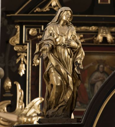 Zdjęcie nr 1: Matka Boska ukazana jest w pozycji stojącej, zwrócona lekko w stronę krzyża, w kontrapoście, ze zgiętą prawą nogą. Głowa delikatnie pochylona w lewo. Twarz Marii owalna, z pełnymi policzkami, zamkniętymi, wąskimi ale pełnymi wargami, prostym, szerokim i małym nosem. Oczy o lekko opadających zewnętrznych końcach zamknięte, nad nimi rysują się delikatne brwi. Na głowę i ramiona Marii narzucony jest płaszcz, spod którego wystają pukle pofalowanych włosów. Prawą rękę z otwartą dłonią wyciąga ku przodowi, lewą zaś zgiętą dotyka piersi. Maria jest ubrana w obficie pofałdowaną suknię, przewiązaną w pasie, sięgającą do ziemi. Poły szaty oraz płaszcza układają się w drobne, ale gęste i głębokie fałdy, swobodnie opinające jej sylwetkę. Rzeźba w całości złocona. Na jej plecach wtórnie dodana deska o obrysie postaci z malowanym wizerunkiem Marii widoczną od strony ołtarza głównego. 