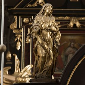 Zdjęcie nr 1: Matka Boska ukazana jest w pozycji stojącej, zwrócona lekko w stronę krzyża, w kontrapoście, ze zgiętą prawą nogą. Głowa delikatnie pochylona w lewo. Twarz Marii owalna, z pełnymi policzkami, zamkniętymi, wąskimi ale pełnymi wargami, prostym, szerokim i małym nosem. Oczy o lekko opadających zewnętrznych końcach zamknięte, nad nimi rysują się delikatne brwi. Na głowę i ramiona Marii narzucony jest płaszcz, spod którego wystają pukle pofalowanych włosów. Prawą rękę z otwartą dłonią wyciąga ku przodowi, lewą zaś zgiętą dotyka piersi. Maria jest ubrana w obficie pofałdowaną suknię, przewiązaną w pasie, sięgającą do ziemi. Poły szaty oraz płaszcza układają się w drobne, ale gęste i głębokie fałdy, swobodnie opinające jej sylwetkę. Rzeźba w całości złocona. Na jej plecach wtórnie dodana deska o obrysie postaci z malowanym wizerunkiem Marii widoczną od strony ołtarza głównego. 