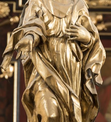 Zdjęcie nr 3: Matka Boska ukazana jest w pozycji stojącej, zwrócona lekko w stronę krzyża, w kontrapoście, ze zgiętą prawą nogą. Głowa delikatnie pochylona w lewo. Twarz Marii owalna, z pełnymi policzkami, zamkniętymi, wąskimi ale pełnymi wargami, prostym, szerokim i małym nosem. Oczy o lekko opadających zewnętrznych końcach zamknięte, nad nimi rysują się delikatne brwi. Na głowę i ramiona Marii narzucony jest płaszcz, spod którego wystają pukle pofalowanych włosów. Prawą rękę z otwartą dłonią wyciąga ku przodowi, lewą zaś zgiętą dotyka piersi. Maria jest ubrana w obficie pofałdowaną suknię, przewiązaną w pasie, sięgającą do ziemi. Poły szaty oraz płaszcza układają się w drobne, ale gęste i głębokie fałdy, swobodnie opinające jej sylwetkę. Rzeźba w całości złocona. Na jej plecach wtórnie dodana deska o obrysie postaci z malowanym wizerunkiem Marii widoczną od strony ołtarza głównego. 