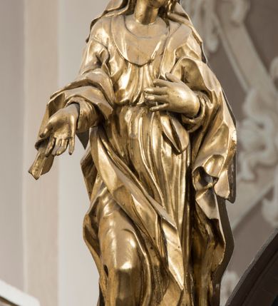 Zdjęcie nr 4: Matka Boska ukazana jest w pozycji stojącej, zwrócona lekko w stronę krzyża, w kontrapoście, ze zgiętą prawą nogą. Głowa delikatnie pochylona w lewo. Twarz Marii owalna, z pełnymi policzkami, zamkniętymi, wąskimi ale pełnymi wargami, prostym, szerokim i małym nosem. Oczy o lekko opadających zewnętrznych końcach zamknięte, nad nimi rysują się delikatne brwi. Na głowę i ramiona Marii narzucony jest płaszcz, spod którego wystają pukle pofalowanych włosów. Prawą rękę z otwartą dłonią wyciąga ku przodowi, lewą zaś zgiętą dotyka piersi. Maria jest ubrana w obficie pofałdowaną suknię, przewiązaną w pasie, sięgającą do ziemi. Poły szaty oraz płaszcza układają się w drobne, ale gęste i głębokie fałdy, swobodnie opinające jej sylwetkę. Rzeźba w całości złocona. Na jej plecach wtórnie dodana deska o obrysie postaci z malowanym wizerunkiem Marii widoczną od strony ołtarza głównego. 