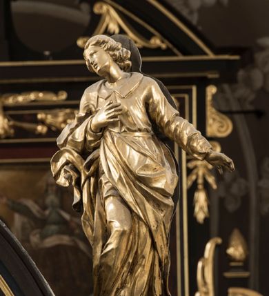 Zdjęcie nr 3: Święty po prawej stronie krzyża, ukazany w pozycji stojącej, w lekkim kontrapoście z ugiętą prawą nogą w kolanie, z głową i górną partią tułowia zwróconą w stronę Ukrzyżowanego, a dolną częścią ciała ukazaną niemal frontalnie. Głowa świętego ukazana w trzech czwartych, lekko przegięta na bok, zwraca się w stronę krzyża. Twarz smukła, z wydatnym podbródkiem i drobnymi ustami lekko rozchylonymi. Mały, prosty, szeroki nos; otwarte, migdałowate oczy od góry zaznaczone delikatnie rysującymi się łukami brwiowymi. Kręcone włosy do ramion układają się w grube pukle, spływają na szyje i zakrywają uszy. Jego prawa ręka jest zgięta w łokciu, przylega do piersi, lewa jest opuszczona i odsunięta od tułowia, z rozłożoną dłonią. Święty Jan jest ubrany w tunikę z kołnierzem i wąskimi rękawami, przewiązaną w pasie i sięgającą do ziemi. Przez prawe ramię przerzucony jest płaszcz, przechodzący pod lewą ręką  z końcem zarzuconym na prawe przedramię. Szaty są mocno pofałdowane, dynamicznie skłębione pod prawą ręką. Rzeźba w całości złocona. Na jej plecach wtórnie dodana deska o obrysie postaci z malowaną wersją świętego widocznego od strony ołtarza głównego.