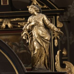 Zdjęcie nr 1: Święty po prawej stronie krzyża, ukazany w pozycji stojącej, w lekkim kontrapoście z ugiętą prawą nogą w kolanie, z głową i górną partią tułowia zwróconą w stronę Ukrzyżowanego, a dolną częścią ciała ukazaną niemal frontalnie. Głowa świętego ukazana w trzech czwartych, lekko przegięta na bok, zwraca się w stronę krzyża. Twarz smukła, z wydatnym podbródkiem i drobnymi ustami lekko rozchylonymi. Mały, prosty, szeroki nos; otwarte, migdałowate oczy od góry zaznaczone delikatnie rysującymi się łukami brwiowymi. Kręcone włosy do ramion układają się w grube pukle, spływają na szyje i zakrywają uszy. Jego prawa ręka jest zgięta w łokciu, przylega do piersi, lewa jest opuszczona i odsunięta od tułowia, z rozłożoną dłonią. Święty Jan jest ubrany w tunikę z kołnierzem i wąskimi rękawami, przewiązaną w pasie i sięgającą do ziemi. Przez prawe ramię przerzucony jest płaszcz, przechodzący pod lewą ręką  z końcem zarzuconym na prawe przedramię. Szaty są mocno pofałdowane, dynamicznie skłębione pod prawą ręką. Rzeźba w całości złocona. Na jej plecach wtórnie dodana deska o obrysie postaci z malowaną wersją świętego widocznego od strony ołtarza głównego.