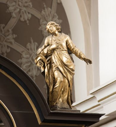 Zdjęcie nr 5: Święty po prawej stronie krzyża, ukazany w pozycji stojącej, w lekkim kontrapoście z ugiętą prawą nogą w kolanie, z głową i górną partią tułowia zwróconą w stronę Ukrzyżowanego, a dolną częścią ciała ukazaną niemal frontalnie. Głowa świętego ukazana w trzech czwartych, lekko przegięta na bok, zwraca się w stronę krzyża. Twarz smukła, z wydatnym podbródkiem i drobnymi ustami lekko rozchylonymi. Mały, prosty, szeroki nos; otwarte, migdałowate oczy od góry zaznaczone delikatnie rysującymi się łukami brwiowymi. Kręcone włosy do ramion układają się w grube pukle, spływają na szyje i zakrywają uszy. Jego prawa ręka jest zgięta w łokciu, przylega do piersi, lewa jest opuszczona i odsunięta od tułowia, z rozłożoną dłonią. Święty Jan jest ubrany w tunikę z kołnierzem i wąskimi rękawami, przewiązaną w pasie i sięgającą do ziemi. Przez prawe ramię przerzucony jest płaszcz, przechodzący pod lewą ręką  z końcem zarzuconym na prawe przedramię. Szaty są mocno pofałdowane, dynamicznie skłębione pod prawą ręką. Rzeźba w całości złocona. Na jej plecach wtórnie dodana deska o obrysie postaci z malowaną wersją świętego widocznego od strony ołtarza głównego.