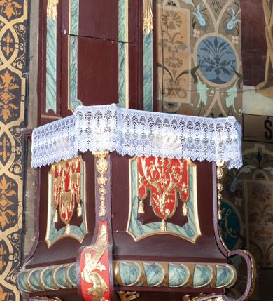 Zdjęcie nr 1: Ambona wisząca, przyścienna, z dostępem od zakrystii. Kosz na planie kwadratu o ściętych narożach, na cokole w formie wałka. U spodu podwieszony odwrócony, ścięty ostrosłup, o wklęsłych bokach, wydzielonych spływami wolutowymi dekorowanymi owocowymi festonami. Pola wypełnione ornamentem roślinnym. Całość zakończona szyszką w kształcie rozety z kulą. Ścianki kosza wydzielone pilastrami w formie wolut, spływających i ujmujących wypukłą cęgą ścięte naroża cokołu; trzony dekorowane owocowymi festonami oraz wstęgą przeplatającą się z ornamentem roślinnym. Podpory dźwigają parapet w formie gzymsu wyłamanego na ich osi. Na ściankach kwadratowe płyciny o wklęsłych narożach i dolnym boku, zwieńczone muszlą. W ich polach sztandary z trzema chwostami z płaskorzeźbioną dekoracją. Frontalny wypełniony wicią utworzoną ze strzępionych palmet i winnego grona; sztandar od strony prezbiterium z trzema obeliskami i podwieszonymi paludamentami w otoczeniu dekoracji roślinnej; sztandar od strony nawy z przedstawieniem trzech sztandarów oraz festonów z podwieszonej tkaniny.  Zaplecek w formie wydłużonego, stojącego prostokąta ujętego lizenami zakończonymi konsolami, dźwigającymi baldachim. Konsole dekorowane akantem z podwieszonymi festonami z tkaniny. W polu zaplecka prostokątna płycina o wklęsłych narożach, w której znajdują się drzwiczki. Baldachim na planie kwadratu o ściętych narożach, w formie niepełnego belkowania, wyłamanego w narożach. Fryz z trzech stron dekorowany kartuszami flankowanymi przez kwiatowe festony, z podwieszonym lambrekinem. U spodu z kwadratową płyciną o wklęsłych narożach. Belkowanie stanowi podstawię dla czterech wolutowych kabłąków podtrzymujących płomienisty wazon, z podwieszoną u spodu szyszką. Na zakończeniach wolut w narożach cztery wazony. 
Struktura polichromowana w kolorze brązowym z czerwonymi oraz marmoryzowanymi w kolorze jasnozielonym detalami; ornamentyka i ramki złocone.