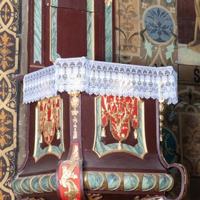 Zdjęcie nr 1: Ambona wisząca, przyścienna, z dostępem od zakrystii. Kosz na planie kwadratu o ściętych narożach, na cokole w formie wałka. U spodu podwieszony odwrócony, ścięty ostrosłup, o wklęsłych bokach, wydzielonych spływami wolutowymi dekorowanymi owocowymi festonami. Pola wypełnione ornamentem roślinnym. Całość zakończona szyszką w kształcie rozety z kulą. Ścianki kosza wydzielone pilastrami w formie wolut, spływających i ujmujących wypukłą cęgą ścięte naroża cokołu; trzony dekorowane owocowymi festonami oraz wstęgą przeplatającą się z ornamentem roślinnym. Podpory dźwigają parapet w formie gzymsu wyłamanego na ich osi. Na ściankach kwadratowe płyciny o wklęsłych narożach i dolnym boku, zwieńczone muszlą. W ich polach sztandary z trzema chwostami z płaskorzeźbioną dekoracją. Frontalny wypełniony wicią utworzoną ze strzępionych palmet i winnego grona; sztandar od strony prezbiterium z trzema obeliskami i podwieszonymi paludamentami w otoczeniu dekoracji roślinnej; sztandar od strony nawy z przedstawieniem trzech sztandarów oraz festonów z podwieszonej tkaniny.  Zaplecek w formie wydłużonego, stojącego prostokąta ujętego lizenami zakończonymi konsolami, dźwigającymi baldachim. Konsole dekorowane akantem z podwieszonymi festonami z tkaniny. W polu zaplecka prostokątna płycina o wklęsłych narożach, w której znajdują się drzwiczki. Baldachim na planie kwadratu o ściętych narożach, w formie niepełnego belkowania, wyłamanego w narożach. Fryz z trzech stron dekorowany kartuszami flankowanymi przez kwiatowe festony, z podwieszonym lambrekinem. U spodu z kwadratową płyciną o wklęsłych narożach. Belkowanie stanowi podstawię dla czterech wolutowych kabłąków podtrzymujących płomienisty wazon, z podwieszoną u spodu szyszką. Na zakończeniach wolut w narożach cztery wazony. 
Struktura polichromowana w kolorze brązowym z czerwonymi oraz marmoryzowanymi w kolorze jasnozielonym detalami; ornamentyka i ramki złocone.