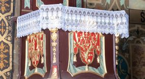 Zdjęcie nr 1: Ambona wisząca, przyścienna, z dostępem od zakrystii. Kosz na planie kwadratu o ściętych narożach, na cokole w formie wałka. U spodu podwieszony odwrócony, ścięty ostrosłup, o wklęsłych bokach, wydzielonych spływami wolutowymi dekorowanymi owocowymi festonami. Pola wypełnione ornamentem roślinnym. Całość zakończona szyszką w kształcie rozety z kulą. Ścianki kosza wydzielone pilastrami w formie wolut, spływających i ujmujących wypukłą cęgą ścięte naroża cokołu; trzony dekorowane owocowymi festonami oraz wstęgą przeplatającą się z ornamentem roślinnym. Podpory dźwigają parapet w formie gzymsu wyłamanego na ich osi. Na ściankach kwadratowe płyciny o wklęsłych narożach i dolnym boku, zwieńczone muszlą. W ich polach sztandary z trzema chwostami z płaskorzeźbioną dekoracją. Frontalny wypełniony wicią utworzoną ze strzępionych palmet i winnego grona; sztandar od strony prezbiterium z trzema obeliskami i podwieszonymi paludamentami w otoczeniu dekoracji roślinnej; sztandar od strony nawy z przedstawieniem trzech sztandarów oraz festonów z podwieszonej tkaniny.  Zaplecek w formie wydłużonego, stojącego prostokąta ujętego lizenami zakończonymi konsolami, dźwigającymi baldachim. Konsole dekorowane akantem z podwieszonymi festonami z tkaniny. W polu zaplecka prostokątna płycina o wklęsłych narożach, w której znajdują się drzwiczki. Baldachim na planie kwadratu o ściętych narożach, w formie niepełnego belkowania, wyłamanego w narożach. Fryz z trzech stron dekorowany kartuszami flankowanymi przez kwiatowe festony, z podwieszonym lambrekinem. U spodu z kwadratową płyciną o wklęsłych narożach. Belkowanie stanowi podstawię dla czterech wolutowych kabłąków podtrzymujących płomienisty wazon, z podwieszoną u spodu szyszką. Na zakończeniach wolut w narożach cztery wazony. 
Struktura polichromowana w kolorze brązowym z czerwonymi oraz marmoryzowanymi w kolorze jasnozielonym detalami; ornamentyka i ramki złocone.