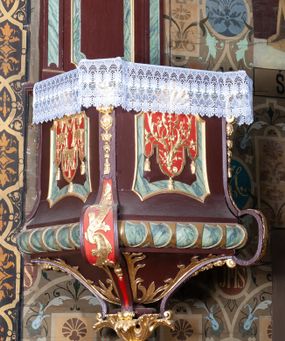 Zdjęcie nr 1: Ambona wisząca, przyścienna, z dostępem od zakrystii. Kosz na planie kwadratu o ściętych narożach, na cokole w formie wałka. U spodu podwieszony odwrócony, ścięty ostrosłup, o wklęsłych bokach, wydzielonych spływami wolutowymi dekorowanymi owocowymi festonami. Pola wypełnione ornamentem roślinnym. Całość zakończona szyszką w kształcie rozety z kulą. Ścianki kosza wydzielone pilastrami w formie wolut, spływających i ujmujących wypukłą cęgą ścięte naroża cokołu; trzony dekorowane owocowymi festonami oraz wstęgą przeplatającą się z ornamentem roślinnym. Podpory dźwigają parapet w formie gzymsu wyłamanego na ich osi. Na ściankach kwadratowe płyciny o wklęsłych narożach i dolnym boku, zwieńczone muszlą. W ich polach sztandary z trzema chwostami z płaskorzeźbioną dekoracją. Frontalny wypełniony wicią utworzoną ze strzępionych palmet i winnego grona; sztandar od strony prezbiterium z trzema obeliskami i podwieszonymi paludamentami w otoczeniu dekoracji roślinnej; sztandar od strony nawy z przedstawieniem trzech sztandarów oraz festonów z podwieszonej tkaniny.  Zaplecek w formie wydłużonego, stojącego prostokąta ujętego lizenami zakończonymi konsolami, dźwigającymi baldachim. Konsole dekorowane akantem z podwieszonymi festonami z tkaniny. W polu zaplecka prostokątna płycina o wklęsłych narożach, w której znajdują się drzwiczki. Baldachim na planie kwadratu o ściętych narożach, w formie niepełnego belkowania, wyłamanego w narożach. Fryz z trzech stron dekorowany kartuszami flankowanymi przez kwiatowe festony, z podwieszonym lambrekinem. U spodu z kwadratową płyciną o wklęsłych narożach. Belkowanie stanowi podstawię dla czterech wolutowych kabłąków podtrzymujących płomienisty wazon, z podwieszoną u spodu szyszką. Na zakończeniach wolut w narożach cztery wazony. 
Struktura polichromowana w kolorze brązowym z czerwonymi oraz marmoryzowanymi w kolorze jasnozielonym detalami; ornamentyka i ramki złocone.