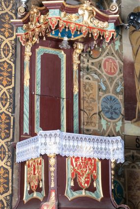 Zdjęcie nr 1: Ambona wisząca, przyścienna, z dostępem od zakrystii. Kosz na planie kwadratu o ściętych narożach, na cokole w formie wałka. U spodu podwieszony odwrócony, ścięty ostrosłup, o wklęsłych bokach, wydzielonych spływami wolutowymi dekorowanymi owocowymi festonami. Pola wypełnione ornamentem roślinnym. Całość zakończona szyszką w kształcie rozety z kulą. Ścianki kosza wydzielone pilastrami w formie wolut, spływających i ujmujących wypukłą cęgą ścięte naroża cokołu; trzony dekorowane owocowymi festonami oraz wstęgą przeplatającą się z ornamentem roślinnym. Podpory dźwigają parapet w formie gzymsu wyłamanego na ich osi. Na ściankach kwadratowe płyciny o wklęsłych narożach i dolnym boku, zwieńczone muszlą. W ich polach sztandary z trzema chwostami z płaskorzeźbioną dekoracją. Frontalny wypełniony wicią utworzoną ze strzępionych palmet i winnego grona; sztandar od strony prezbiterium z trzema obeliskami i podwieszonymi paludamentami w otoczeniu dekoracji roślinnej; sztandar od strony nawy z przedstawieniem trzech sztandarów oraz festonów z podwieszonej tkaniny.  Zaplecek w formie wydłużonego, stojącego prostokąta ujętego lizenami zakończonymi konsolami, dźwigającymi baldachim. Konsole dekorowane akantem z podwieszonymi festonami z tkaniny. W polu zaplecka prostokątna płycina o wklęsłych narożach, w której znajdują się drzwiczki. Baldachim na planie kwadratu o ściętych narożach, w formie niepełnego belkowania, wyłamanego w narożach. Fryz z trzech stron dekorowany kartuszami flankowanymi przez kwiatowe festony, z podwieszonym lambrekinem. U spodu z kwadratową płyciną o wklęsłych narożach. Belkowanie stanowi podstawię dla czterech wolutowych kabłąków podtrzymujących płomienisty wazon, z podwieszoną u spodu szyszką. Na zakończeniach wolut w narożach cztery wazony. 
Struktura polichromowana w kolorze brązowym z czerwonymi oraz marmoryzowanymi w kolorze jasnozielonym detalami; ornamentyka i ramki złocone.