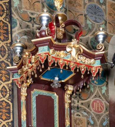 Zdjęcie nr 2: Ambona wisząca, przyścienna, z dostępem od zakrystii. Kosz na planie kwadratu o ściętych narożach, na cokole w formie wałka. U spodu podwieszony odwrócony, ścięty ostrosłup, o wklęsłych bokach, wydzielonych spływami wolutowymi dekorowanymi owocowymi festonami. Pola wypełnione ornamentem roślinnym. Całość zakończona szyszką w kształcie rozety z kulą. Ścianki kosza wydzielone pilastrami w formie wolut, spływających i ujmujących wypukłą cęgą ścięte naroża cokołu; trzony dekorowane owocowymi festonami oraz wstęgą przeplatającą się z ornamentem roślinnym. Podpory dźwigają parapet w formie gzymsu wyłamanego na ich osi. Na ściankach kwadratowe płyciny o wklęsłych narożach i dolnym boku, zwieńczone muszlą. W ich polach sztandary z trzema chwostami z płaskorzeźbioną dekoracją. Frontalny wypełniony wicią utworzoną ze strzępionych palmet i winnego grona; sztandar od strony prezbiterium z trzema obeliskami i podwieszonymi paludamentami w otoczeniu dekoracji roślinnej; sztandar od strony nawy z przedstawieniem trzech sztandarów oraz festonów z podwieszonej tkaniny.  Zaplecek w formie wydłużonego, stojącego prostokąta ujętego lizenami zakończonymi konsolami, dźwigającymi baldachim. Konsole dekorowane akantem z podwieszonymi festonami z tkaniny. W polu zaplecka prostokątna płycina o wklęsłych narożach, w której znajdują się drzwiczki. Baldachim na planie kwadratu o ściętych narożach, w formie niepełnego belkowania, wyłamanego w narożach. Fryz z trzech stron dekorowany kartuszami flankowanymi przez kwiatowe festony, z podwieszonym lambrekinem. U spodu z kwadratową płyciną o wklęsłych narożach. Belkowanie stanowi podstawię dla czterech wolutowych kabłąków podtrzymujących płomienisty wazon, z podwieszoną u spodu szyszką. Na zakończeniach wolut w narożach cztery wazony. 
Struktura polichromowana w kolorze brązowym z czerwonymi oraz marmoryzowanymi w kolorze jasnozielonym detalami; ornamentyka i ramki złocone.