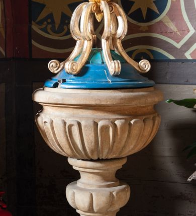 Zdjęcie nr 1: Chrzcielnica w typie kielichowym na płaskiej podstawie na planie koła, osadzona na niewysokim, ośmiobocznym postumencie. Stopa niewielka, ujęta półwałkiem. Spłaszczony, puklowany nodus trzonu ujęty parą talerzyków. Czara półkulistą o powierzchni imitującej puklowanie z wklęską i zaoblonym brzegiem. Stożkowata pokrywa zwieńczona sześcioma kabłąkami w formie spływów wolutowych, spiętymi ujętą dołem sterczynką zamkniętą kulą z krzyżem. Struktura kielicha chrzcielnicy utrzymana w kolorze kamienia, pokrywa polichromowana w kolorze niebieskim i turkusowym, zwieńczenie złocono-srebrzone.  