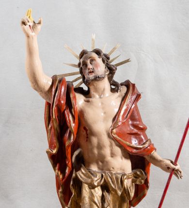 Zdjęcie nr 1: Rzeźba całopostaciowa, pełnoplastyczna, ustawiona na cokole w formie obłoku. Sylwetka Jezusa zwrócona na wprost, w wydatnym kontrapoście, z prawą nogą wspartą na leżącej na podstawie czaszce. Głowa Jezusa uniesiona, skierowana delikatnie w prawo. Twarz pociągła, o kwadratowym zarysie szczęki, o głęboko osadzonych oczach z drążonymi kącikami, zwróconych ku górze; prostym, długim nosie, rozchylonych ustach; okolona krótką kręconą brodą, układającą się w dwa pukle. Włosy długie, gęste, kręcone, układające się w wijące pasma, opadające na ramiona. Jezus w lewej ręce trzyma czerwony sztandar na drzewcu zakończonym krzyżem, prawą unosi wysoko w geście błogosławieństwa. Ciało o delikatnie zarysowanej muskulaturze z zaznaczonymi śladami męki. W partii bioder przepasany złoconym perizonium przewiązanym sznurem, sięgającym połowy uda i odsłaniającym lewe biodro; tkanina drapowana w grube fałdy, marszczona, z wywiniętym brzegiem. Na ramiona narzucony czerwony płaszcz opadający na plecy, drapowany w proste fałdy, z zawiniętym brzegiem. Głowę ujmuje gloria promienista.