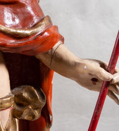 Zdjęcie nr 7: Rzeźba całopostaciowa, pełnoplastyczna, ustawiona na cokole w formie obłoku. Sylwetka Jezusa zwrócona na wprost, w wydatnym kontrapoście, z prawą nogą wspartą na leżącej na podstawie czaszce. Głowa Jezusa uniesiona, skierowana delikatnie w prawo. Twarz pociągła, o kwadratowym zarysie szczęki, o głęboko osadzonych oczach z drążonymi kącikami, zwróconych ku górze; prostym, długim nosie, rozchylonych ustach; okolona krótką kręconą brodą, układającą się w dwa pukle. Włosy długie, gęste, kręcone, układające się w wijące pasma, opadające na ramiona. Jezus w lewej ręce trzyma czerwony sztandar na drzewcu zakończonym krzyżem, prawą unosi wysoko w geście błogosławieństwa. Ciało o delikatnie zarysowanej muskulaturze z zaznaczonymi śladami męki. W partii bioder przepasany złoconym perizonium przewiązanym sznurem, sięgającym połowy uda i odsłaniającym lewe biodro; tkanina drapowana w grube fałdy, marszczona, z wywiniętym brzegiem. Na ramiona narzucony czerwony płaszcz opadający na plecy, drapowany w proste fałdy, z zawiniętym brzegiem. Głowę ujmuje gloria promienista.