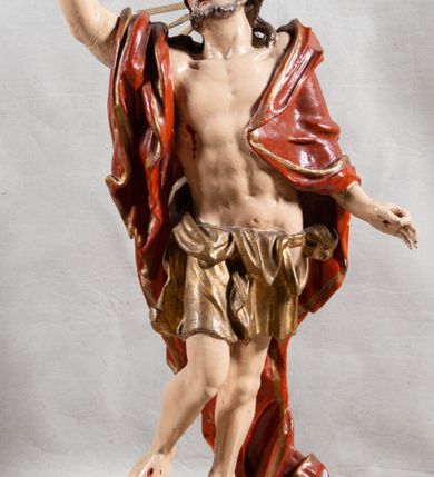 Zdjęcie nr 2: Rzeźba całopostaciowa, pełnoplastyczna, ustawiona na cokole w formie obłoku. Sylwetka Jezusa zwrócona na wprost, w wydatnym kontrapoście, z prawą nogą wspartą na leżącej na podstawie czaszce. Głowa Jezusa uniesiona, skierowana delikatnie w prawo. Twarz pociągła, o kwadratowym zarysie szczęki, o głęboko osadzonych oczach z drążonymi kącikami, zwróconych ku górze; prostym, długim nosie, rozchylonych ustach; okolona krótką kręconą brodą, układającą się w dwa pukle. Włosy długie, gęste, kręcone, układające się w wijące pasma, opadające na ramiona. Jezus w lewej ręce trzyma czerwony sztandar na drzewcu zakończonym krzyżem, prawą unosi wysoko w geście błogosławieństwa. Ciało o delikatnie zarysowanej muskulaturze z zaznaczonymi śladami męki. W partii bioder przepasany złoconym perizonium przewiązanym sznurem, sięgającym połowy uda i odsłaniającym lewe biodro; tkanina drapowana w grube fałdy, marszczona, z wywiniętym brzegiem. Na ramiona narzucony czerwony płaszcz opadający na plecy, drapowany w proste fałdy, z zawiniętym brzegiem. Głowę ujmuje gloria promienista.