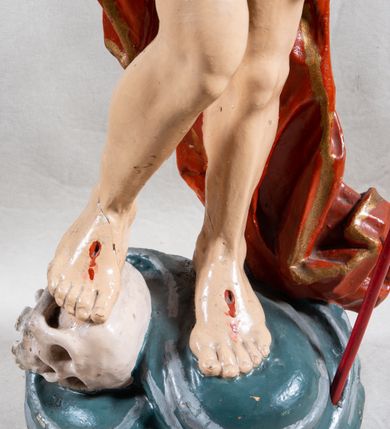 Zdjęcie nr 8: Rzeźba całopostaciowa, pełnoplastyczna, ustawiona na cokole w formie obłoku. Sylwetka Jezusa zwrócona na wprost, w wydatnym kontrapoście, z prawą nogą wspartą na leżącej na podstawie czaszce. Głowa Jezusa uniesiona, skierowana delikatnie w prawo. Twarz pociągła, o kwadratowym zarysie szczęki, o głęboko osadzonych oczach z drążonymi kącikami, zwróconych ku górze; prostym, długim nosie, rozchylonych ustach; okolona krótką kręconą brodą, układającą się w dwa pukle. Włosy długie, gęste, kręcone, układające się w wijące pasma, opadające na ramiona. Jezus w lewej ręce trzyma czerwony sztandar na drzewcu zakończonym krzyżem, prawą unosi wysoko w geście błogosławieństwa. Ciało o delikatnie zarysowanej muskulaturze z zaznaczonymi śladami męki. W partii bioder przepasany złoconym perizonium przewiązanym sznurem, sięgającym połowy uda i odsłaniającym lewe biodro; tkanina drapowana w grube fałdy, marszczona, z wywiniętym brzegiem. Na ramiona narzucony czerwony płaszcz opadający na plecy, drapowany w proste fałdy, z zawiniętym brzegiem. Głowę ujmuje gloria promienista.