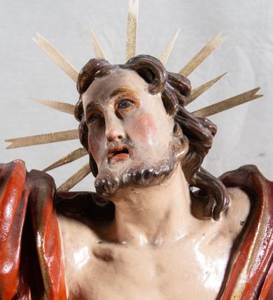 Zdjęcie nr 4: Rzeźba całopostaciowa, pełnoplastyczna, ustawiona na cokole w formie obłoku. Sylwetka Jezusa zwrócona na wprost, w wydatnym kontrapoście, z prawą nogą wspartą na leżącej na podstawie czaszce. Głowa Jezusa uniesiona, skierowana delikatnie w prawo. Twarz pociągła, o kwadratowym zarysie szczęki, o głęboko osadzonych oczach z drążonymi kącikami, zwróconych ku górze; prostym, długim nosie, rozchylonych ustach; okolona krótką kręconą brodą, układającą się w dwa pukle. Włosy długie, gęste, kręcone, układające się w wijące pasma, opadające na ramiona. Jezus w lewej ręce trzyma czerwony sztandar na drzewcu zakończonym krzyżem, prawą unosi wysoko w geście błogosławieństwa. Ciało o delikatnie zarysowanej muskulaturze z zaznaczonymi śladami męki. W partii bioder przepasany złoconym perizonium przewiązanym sznurem, sięgającym połowy uda i odsłaniającym lewe biodro; tkanina drapowana w grube fałdy, marszczona, z wywiniętym brzegiem. Na ramiona narzucony czerwony płaszcz opadający na plecy, drapowany w proste fałdy, z zawiniętym brzegiem. Głowę ujmuje gloria promienista.