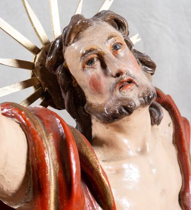 Zdjęcie nr 5: Rzeźba całopostaciowa, pełnoplastyczna, ustawiona na cokole w formie obłoku. Sylwetka Jezusa zwrócona na wprost, w wydatnym kontrapoście, z prawą nogą wspartą na leżącej na podstawie czaszce. Głowa Jezusa uniesiona, skierowana delikatnie w prawo. Twarz pociągła, o kwadratowym zarysie szczęki, o głęboko osadzonych oczach z drążonymi kącikami, zwróconych ku górze; prostym, długim nosie, rozchylonych ustach; okolona krótką kręconą brodą, układającą się w dwa pukle. Włosy długie, gęste, kręcone, układające się w wijące pasma, opadające na ramiona. Jezus w lewej ręce trzyma czerwony sztandar na drzewcu zakończonym krzyżem, prawą unosi wysoko w geście błogosławieństwa. Ciało o delikatnie zarysowanej muskulaturze z zaznaczonymi śladami męki. W partii bioder przepasany złoconym perizonium przewiązanym sznurem, sięgającym połowy uda i odsłaniającym lewe biodro; tkanina drapowana w grube fałdy, marszczona, z wywiniętym brzegiem. Na ramiona narzucony czerwony płaszcz opadający na plecy, drapowany w proste fałdy, z zawiniętym brzegiem. Głowę ujmuje gloria promienista.