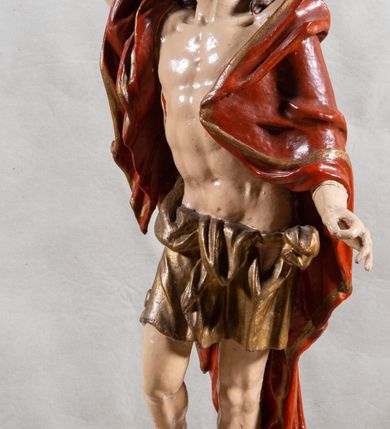 Zdjęcie nr 13: Rzeźba całopostaciowa, pełnoplastyczna, ustawiona na cokole w formie obłoku. Sylwetka Jezusa zwrócona na wprost, w wydatnym kontrapoście, z prawą nogą wspartą na leżącej na podstawie czaszce. Głowa Jezusa uniesiona, skierowana delikatnie w prawo. Twarz pociągła, o kwadratowym zarysie szczęki, o głęboko osadzonych oczach z drążonymi kącikami, zwróconych ku górze; prostym, długim nosie, rozchylonych ustach; okolona krótką kręconą brodą, układającą się w dwa pukle. Włosy długie, gęste, kręcone, układające się w wijące pasma, opadające na ramiona. Jezus w lewej ręce trzyma czerwony sztandar na drzewcu zakończonym krzyżem, prawą unosi wysoko w geście błogosławieństwa. Ciało o delikatnie zarysowanej muskulaturze z zaznaczonymi śladami męki. W partii bioder przepasany złoconym perizonium przewiązanym sznurem, sięgającym połowy uda i odsłaniającym lewe biodro; tkanina drapowana w grube fałdy, marszczona, z wywiniętym brzegiem. Na ramiona narzucony czerwony płaszcz opadający na plecy, drapowany w proste fałdy, z zawiniętym brzegiem. Głowę ujmuje gloria promienista.