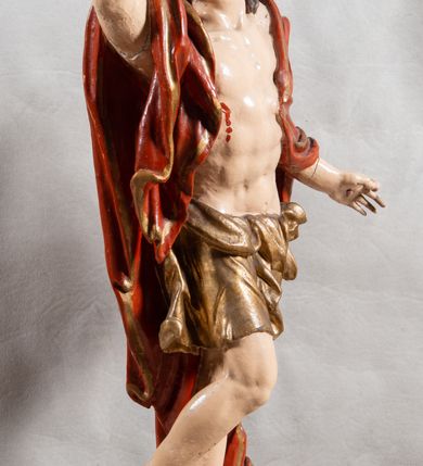 Zdjęcie nr 10: Rzeźba całopostaciowa, pełnoplastyczna, ustawiona na cokole w formie obłoku. Sylwetka Jezusa zwrócona na wprost, w wydatnym kontrapoście, z prawą nogą wspartą na leżącej na podstawie czaszce. Głowa Jezusa uniesiona, skierowana delikatnie w prawo. Twarz pociągła, o kwadratowym zarysie szczęki, o głęboko osadzonych oczach z drążonymi kącikami, zwróconych ku górze; prostym, długim nosie, rozchylonych ustach; okolona krótką kręconą brodą, układającą się w dwa pukle. Włosy długie, gęste, kręcone, układające się w wijące pasma, opadające na ramiona. Jezus w lewej ręce trzyma czerwony sztandar na drzewcu zakończonym krzyżem, prawą unosi wysoko w geście błogosławieństwa. Ciało o delikatnie zarysowanej muskulaturze z zaznaczonymi śladami męki. W partii bioder przepasany złoconym perizonium przewiązanym sznurem, sięgającym połowy uda i odsłaniającym lewe biodro; tkanina drapowana w grube fałdy, marszczona, z wywiniętym brzegiem. Na ramiona narzucony czerwony płaszcz opadający na plecy, drapowany w proste fałdy, z zawiniętym brzegiem. Głowę ujmuje gloria promienista.