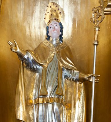 Zdjęcie nr 1: Święty ukazany frontalnie, w kontrapoście, o lekko odchylonej sylwetce, z rękami rozłożonymi na boki. Prawa dłoń w geście błogosławieństwa, w lewej trzyma pastorał o krzywaśni dekorowanej akantem. Twarz ma kwadratową, o dużych, migdałowatych oczach, krótkim nosie i małych ustach, okoloną kręconymi, siwymi włosami sięgającymi ramion. Na głowie ma złoconą infułę z imitacją kameryzacji. Ubrany jest w srebrzoną albę, srebrzoną rokietę o złoconym brzegu, złoconą stułę oraz złoconą kapę o płaskorzeźbionej bordiurze, zapiętą na piersi klamrą. Na dłoniach ma złocone rękawiczki. Polichromia ciała naturalistyczna. 