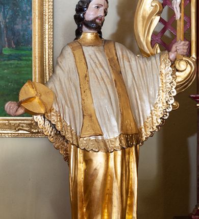 Zdjęcie nr 2: Figura ustawiona na niskiej podstawie. Święty ukazany frontalnie, z głową skierowaną w lewo, w kontrapoście, z rękami rozłożonymi na boki. W lewej dłoni trzyma krucyfiks, w prawej złocony biret. Twarz ma kwadratową, o migdałowatych oczach, krótkim, dużym nosie i wąskich ustach, okoloną krótką brodą oraz kręconymi włosami sięgającymi ramion. Ubrany jest w złoconą albę, biały ornat, lamowany złoconą koronką oraz złoconą stułę. Polichromia ciała naturalistyczna.