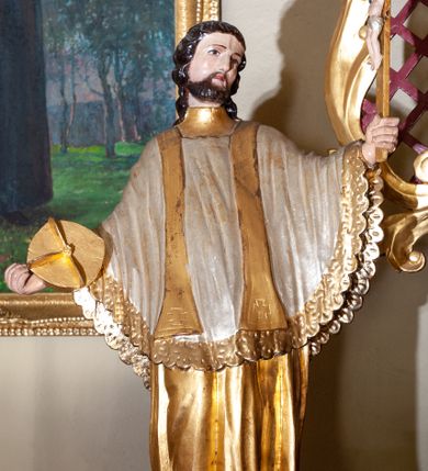 Zdjęcie nr 1: Figura ustawiona na niskiej podstawie. Święty ukazany frontalnie, z głową skierowaną w lewo, w kontrapoście, z rękami rozłożonymi na boki. W lewej dłoni trzyma krucyfiks, w prawej złocony biret. Twarz ma kwadratową, o migdałowatych oczach, krótkim, dużym nosie i wąskich ustach, okoloną krótką brodą oraz kręconymi włosami sięgającymi ramion. Ubrany jest w złoconą albę, biały ornat, lamowany złoconą koronką oraz złoconą stułę. Polichromia ciała naturalistyczna.