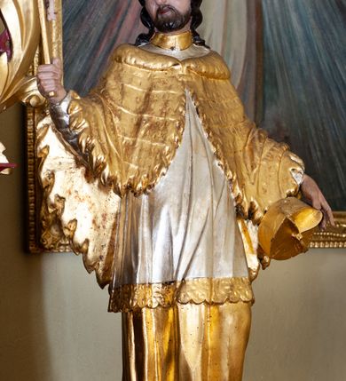 Zdjęcie nr 2: Figura ustawiona na niskiej podstawie. Święty ukazany frontalnie, z głową skierowaną w prawo, w kontrapoście. W prawej, ugiętej i uniesionej ręce trzyma krucyfiks, lewą przytrzymuje przy boku biret. Twarz ma kwadratową, o migdałowatych oczach, krótkim, dużym nosie i wąskich ustach, okoloną krótką brodą i kręconymi włosami sięgającymi ramion. Ubrany jest w złoconą albę, srebrzoną komże ze złoconą koronką przy brzegu oraz złoconą almucję zapiętą pod szyją. Polichromia ciała naturalistyczna. 