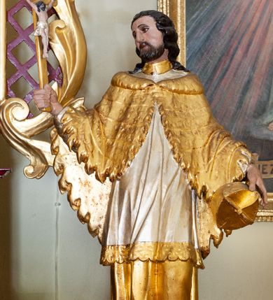 Zdjęcie nr 1: Figura ustawiona na niskiej podstawie. Święty ukazany frontalnie, z głową skierowaną w prawo, w kontrapoście. W prawej, ugiętej i uniesionej ręce trzyma krucyfiks, lewą przytrzymuje przy boku biret. Twarz ma kwadratową, o migdałowatych oczach, krótkim, dużym nosie i wąskich ustach, okoloną krótką brodą i kręconymi włosami sięgającymi ramion. Ubrany jest w złoconą albę, srebrzoną komże ze złoconą koronką przy brzegu oraz złoconą almucję zapiętą pod szyją. Polichromia ciała naturalistyczna. 