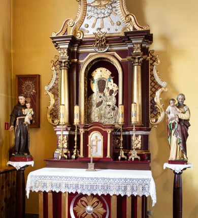 Zdjęcie nr 2: Nastawa architektoniczna, jednoosiowa, jednokondygnacyjna ze zwieńczeniem, ustawiona na cokole z wyodrębnionymi prostopadłościanami. Ołtarz drewniany, prostopadłościenny, z drewnianą mensą. Antepedium flankowane dwiema parami pilastrów, z wyodrębnioną kwadratową płyciną z medalionem z rozetą, ujęte parą pilastrów. Cokół dekorowany rozetami i akantem. Nastawa flankowana dwiema kolumnami o kapitelach kompozytowych, dźwigających pełne belkowanie wyłamane na ich osi oraz częściowo przerwane nad polem środkowym, z rozetami w partii fryzu oraz fragmenty przerwanego, wolutowego przyczółka. W polu głównym wnęka w formie stojącego prostokąta zamkniętego łukiem półkolistym, ujęta profilowaną ramą, z obrazem Matki Boskiej Częstochowskiej. Nad obrazem monogram Marii na sierpie księżyca. Kondygnacja ujęta uszami utworzonymi z akantu, kratki regencyjnej i karbowanej wstęgi. Zwieńczenie w formie zbliżonej do trapezu, zamkniętego dwiema stykającymi się wolutami z muszlą, ujętego spływami wolutowymi, całość obwiedziona karbowaną wstęgą. W polu płaskorzeźbione przedstawienie półpostaci Boga Ojca pośród obłoków, w glorii promienistej, w otoczeniu z gwiazd. Struktura polichromowana w kolorze ciemnoczerwonym, ze złoconymi kolumnami, profilami i ornamentyką oraz srebrzonym polem zwieńczenia i detalami. 