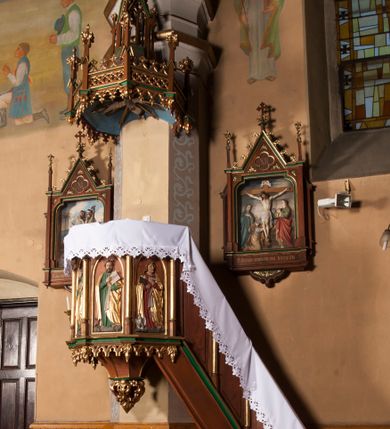 Zdjęcie nr 1: Ambona wisząca, przyścienna, składająca się z kosza, baldachimu i schodów z balustradą. Kosz na planie ośmioboku, podzielony na pola gładkimi, złoconymi kolumnami podtrzymującymi gzyms i parapet. W polach płyciny w formie stojącego prostokąta zamkniętego łukiem ostrym. W płycinach całopostaciowe figury ewangelistów – od strony schodów – św. Jan, św. Łukasz, św. Marek i św. Mateusz. Każdy z nich ubrany w długą suknię, z narzuconym złotym płaszczem, trzyma w rękach otwartą księgę i ma przy nodze właściwy atrybut. Wzdłuż ramy dolnej ażurowy ornament maswerkowy. Do podstawy podwieszona szyszka złożona z liści akantu. Baldachim na planie części ośmioboku o architektonicznym zwieńczeniu. Daszek zdobiony ażurowymi ornamentami maswerkowymi, z pinaklami zwieńczonymi kwiatonami na narożach. Szczyt w formie ażurowej wieży zwieńczonej kwiatonem. W podniebieniu baldachimu wyobrażenie gołębicy Ducha Świętego na tle glorii promienistej. Schody dostawione od strony wschodniej, dwukrotnie załamane, z czterema gładkimi płycinami oddzielonymi złoconymi kolumnami.