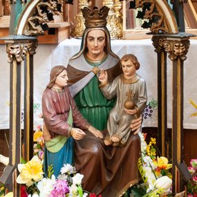 Zdjęcie nr 1: Feretron z przedstawieniem św. Anny Samotrzeć pod neogotyckim, ostrołukowym baldachimem. W centrum siedzi św. Anna, która prawą ręką obejmuje stojącą po jej prawej stronie Marię, a na lewym kolanie trzyma małego Chrystusa. Anna ma owalną twarz o wyrazistych rysach, małe oczy, prosty nos i wąskie usta; spogląda przed siebie. Jest ubrana w zieloną, przepasaną suknię oraz brązowy płaszcz podbity białym suknem. Na głowie ma obszyty złotem welon w kolorze sukni i otwartą koronę. Maria stoi zwrócona w stronę Chrystusa, wyciąga obie ręce przed siebie. Ma owalną twarz o delikatnych rysach i jasne, spływające na plecy włosy. Jest ubrana w czerwoną, przepasaną suknię oraz podbity zielenią niebieski płaszcz, którzy przechodzi pod lewą ręką i wokół prawego biodra. Jezus siedzi, unosi prawą rękę w geście błogosławieństwa, w lewej trzyma jabłko królewskie. Ma okrągłą twarz o delikatnych rysach i jasne włosy opadające na uszy. Jest ubrany w białą sukienkę obszytą złotem pod szyją, na rękawach i przy dolnej krawędzi. Polichromia postaci w partiach ciała naturalistyczna. Grupa siedzi pod baldachimem opartym na czterech smukłych  brązowo-złotych kolumnach o kapitelach utworzonych z liści akantu. Na nich wznosi się ostrołukowy, zielony daszek zdobiony od dołu ażurową koronką, zwieńczony szczytem przeprutym trójliściem i zdobiony na narożach pinaklami zakończonymi kwiatonami. Detale i ornamenty złocone. Podstawa prostopadłościenna.