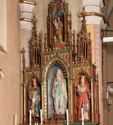 Zdjęcie nr 1: Ołtarz przyścienny, architektoniczny, trójosiowy, jednokondygnacyjny ze zwieńczeniem. Mensa skrzyniowa, prostopadłościenna. Antepedium z trzema prostokątnymi płycinami. W środkowej monogram Marii, boczne wypełnione symetrycznym ornamentem roślinnym. Predella wysoka, dekorowana fryzem arkadkowym, ostrołukowym. W pierwszej kondygnacji retabulum trzy nisze prostokątne zamknięte od góry wimpergą na tle szczytu. Środkowa nisza wyższa i szersza od bocznych, rama dekorowana wyschniętą wicią roślinną, wszystkie zdobione ażurowym ornamentem ostrołukowym i żabkami, szczyty wypełniają pola zamknięte łukiem trójlistnym, ostrym.  Boczne nisze zwieńczone ustawionymi skośnie ażurowymi kapliczkami z pinaklami, zdobionymi żabkami i kwiatonami. W środkowej niszy figura Matki Boskiej z Lourdes, w bocznych św. Kazimierza i św. Agnieszki. W zwieńczeniu ażurowa kapliczka powtarzająca kształt kapliczek wieńczących osie boczne pierwszej kondygnacji z niszą, w której figura św. Józefa z Dzieciątkiem. 
Struktura brązowa, niektóre detale malowane na zielono, ornamenty, profile i kolumny złocone. 