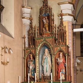 Zdjęcie nr 1: Ołtarz przyścienny, architektoniczny, trójosiowy, jednokondygnacyjny ze zwieńczeniem. Mensa skrzyniowa, prostopadłościenna. Antepedium z trzema prostokątnymi płycinami. W środkowej monogram Marii, boczne wypełnione symetrycznym ornamentem roślinnym. Predella wysoka, dekorowana fryzem arkadkowym, ostrołukowym. W pierwszej kondygnacji retabulum trzy nisze prostokątne zamknięte od góry wimpergą na tle szczytu. Środkowa nisza wyższa i szersza od bocznych, rama dekorowana wyschniętą wicią roślinną, wszystkie zdobione ażurowym ornamentem ostrołukowym i żabkami, szczyty wypełniają pola zamknięte łukiem trójlistnym, ostrym.  Boczne nisze zwieńczone ustawionymi skośnie ażurowymi kapliczkami z pinaklami, zdobionymi żabkami i kwiatonami. W środkowej niszy figura Matki Boskiej z Lourdes, w bocznych św. Kazimierza i św. Agnieszki. W zwieńczeniu ażurowa kapliczka powtarzająca kształt kapliczek wieńczących osie boczne pierwszej kondygnacji z niszą, w której figura św. Józefa z Dzieciątkiem. 
Struktura brązowa, niektóre detale malowane na zielono, ornamenty, profile i kolumny złocone. 