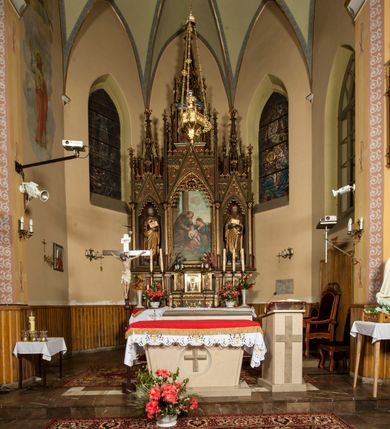 Zdjęcie nr 2: Nastawa ołtarzowa architektoniczna, trójosiowa, jednokondygnacyjna ze zwieńczeniem i predellą. Mensa skrzyniowa, prostopadłościenna. Antepedium z profilowaną płyciną, w centrum medalion z monogramem IHS i gorejącym sercem w glorii. Medalion ujęty profilowanymi płycinami o krawędziach wklęsłych od strony medaliony. Predella wysoka trójdzielna, boczne płyciny dekorowane fryzem arkadkowym ostrołukowym, przed środkową ustawione tabernakulum. W pierwszej kondygnacji retabulum trzy nisze prostokątne zamknięte od góry wimpergą. Środkowa nisza wyższa i szersza od bocznych, rama dekorowana astwerkiem, zwieńczenie łuku wypełnia ażurowy ornament ostrołukowy złączony gałązkami wyschniętej wici. Boczne nisze zdobione ażurowym ornamentem ostrołukowym i żabkami, szczyty wypełniają pola z łukiem trójlistnym ostrym oraz pas dekoracji z rombów wypełnionych motywem czteroliścia. Boczne nisze zwieńczone ustawionymi skośnie ażurowymi kapliczkami z pinaklami, zdobionymi żabkami i kwiatonami. W środkowej niszy obraz z przedstawieniem św. Anny z Marią i Jezusem, w bocznych figury św. Piotra i św. Pawła. W zwieńczeniu bardzo rozbudowana ażurowa kapliczka z niszą z rzeźbionym przedstawieniem Trójcy Świętej. Oprawa tabernakulum trójosiowa, na wysokim cokole zamkniętym wydatnym gzymsem oraz fryzem z astwerkiem; osie boczne cofnięte. Cokół dekorowany esownicami z akantem, boczne różami. Na osi środkowej tabernakulum o groszkowanych drzwiczkach w formie stojącego prostokąta z aplikowanymi kielichem z hostią oraz flankującą go winną latoroślą. Na osiach bocznych blendy z płaskorzeźbionym motywem roślinnym. 
Struktura brązowa, niektóre detale malowane na zielono, ornamenty, profile i kolumny złocone; mensa malowana na biało, ze złoconymi profilami. 