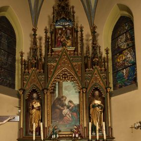 Zdjęcie nr 1: Nastawa ołtarzowa architektoniczna, trójosiowa, jednokondygnacyjna ze zwieńczeniem i predellą. Mensa skrzyniowa, prostopadłościenna. Antepedium z profilowaną płyciną, w centrum medalion z monogramem IHS i gorejącym sercem w glorii. Medalion ujęty profilowanymi płycinami o krawędziach wklęsłych od strony medaliony. Predella wysoka trójdzielna, boczne płyciny dekorowane fryzem arkadkowym ostrołukowym, przed środkową ustawione tabernakulum. W pierwszej kondygnacji retabulum trzy nisze prostokątne zamknięte od góry wimpergą. Środkowa nisza wyższa i szersza od bocznych, rama dekorowana astwerkiem, zwieńczenie łuku wypełnia ażurowy ornament ostrołukowy złączony gałązkami wyschniętej wici. Boczne nisze zdobione ażurowym ornamentem ostrołukowym i żabkami, szczyty wypełniają pola z łukiem trójlistnym ostrym oraz pas dekoracji z rombów wypełnionych motywem czteroliścia. Boczne nisze zwieńczone ustawionymi skośnie ażurowymi kapliczkami z pinaklami, zdobionymi żabkami i kwiatonami. W środkowej niszy obraz z przedstawieniem św. Anny z Marią i Jezusem, w bocznych figury św. Piotra i św. Pawła. W zwieńczeniu bardzo rozbudowana ażurowa kapliczka z niszą z rzeźbionym przedstawieniem Trójcy Świętej. Oprawa tabernakulum trójosiowa, na wysokim cokole zamkniętym wydatnym gzymsem oraz fryzem z astwerkiem; osie boczne cofnięte. Cokół dekorowany esownicami z akantem, boczne różami. Na osi środkowej tabernakulum o groszkowanych drzwiczkach w formie stojącego prostokąta z aplikowanymi kielichem z hostią oraz flankującą go winną latoroślą. Na osiach bocznych blendy z płaskorzeźbionym motywem roślinnym. 
Struktura brązowa, niektóre detale malowane na zielono, ornamenty, profile i kolumny złocone; mensa malowana na biało, ze złoconymi profilami. 