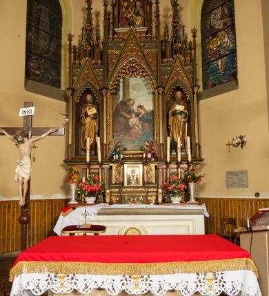 Zdjęcie nr 4: Nastawa ołtarzowa architektoniczna, trójosiowa, jednokondygnacyjna ze zwieńczeniem i predellą. Mensa skrzyniowa, prostopadłościenna. Antepedium z profilowaną płyciną, w centrum medalion z monogramem IHS i gorejącym sercem w glorii. Medalion ujęty profilowanymi płycinami o krawędziach wklęsłych od strony medaliony. Predella wysoka trójdzielna, boczne płyciny dekorowane fryzem arkadkowym ostrołukowym, przed środkową ustawione tabernakulum. W pierwszej kondygnacji retabulum trzy nisze prostokątne zamknięte od góry wimpergą. Środkowa nisza wyższa i szersza od bocznych, rama dekorowana astwerkiem, zwieńczenie łuku wypełnia ażurowy ornament ostrołukowy złączony gałązkami wyschniętej wici. Boczne nisze zdobione ażurowym ornamentem ostrołukowym i żabkami, szczyty wypełniają pola z łukiem trójlistnym ostrym oraz pas dekoracji z rombów wypełnionych motywem czteroliścia. Boczne nisze zwieńczone ustawionymi skośnie ażurowymi kapliczkami z pinaklami, zdobionymi żabkami i kwiatonami. W środkowej niszy obraz z przedstawieniem św. Anny z Marią i Jezusem, w bocznych figury św. Piotra i św. Pawła. W zwieńczeniu bardzo rozbudowana ażurowa kapliczka z niszą z rzeźbionym przedstawieniem Trójcy Świętej. Oprawa tabernakulum trójosiowa, na wysokim cokole zamkniętym wydatnym gzymsem oraz fryzem z astwerkiem; osie boczne cofnięte. Cokół dekorowany esownicami z akantem, boczne różami. Na osi środkowej tabernakulum o groszkowanych drzwiczkach w formie stojącego prostokąta z aplikowanymi kielichem z hostią oraz flankującą go winną latoroślą. Na osiach bocznych blendy z płaskorzeźbionym motywem roślinnym. 
Struktura brązowa, niektóre detale malowane na zielono, ornamenty, profile i kolumny złocone; mensa malowana na biało, ze złoconymi profilami. 