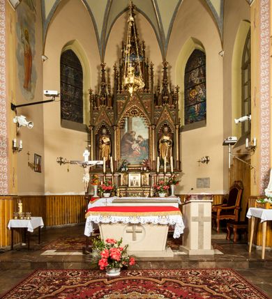 Zdjęcie nr 5: Nastawa ołtarzowa architektoniczna, trójosiowa, jednokondygnacyjna ze zwieńczeniem i predellą. Mensa skrzyniowa, prostopadłościenna. Antepedium z profilowaną płyciną, w centrum medalion z monogramem IHS i gorejącym sercem w glorii. Medalion ujęty profilowanymi płycinami o krawędziach wklęsłych od strony medaliony. Predella wysoka trójdzielna, boczne płyciny dekorowane fryzem arkadkowym ostrołukowym, przed środkową ustawione tabernakulum. W pierwszej kondygnacji retabulum trzy nisze prostokątne zamknięte od góry wimpergą. Środkowa nisza wyższa i szersza od bocznych, rama dekorowana astwerkiem, zwieńczenie łuku wypełnia ażurowy ornament ostrołukowy złączony gałązkami wyschniętej wici. Boczne nisze zdobione ażurowym ornamentem ostrołukowym i żabkami, szczyty wypełniają pola z łukiem trójlistnym ostrym oraz pas dekoracji z rombów wypełnionych motywem czteroliścia. Boczne nisze zwieńczone ustawionymi skośnie ażurowymi kapliczkami z pinaklami, zdobionymi żabkami i kwiatonami. W środkowej niszy obraz z przedstawieniem św. Anny z Marią i Jezusem, w bocznych figury św. Piotra i św. Pawła. W zwieńczeniu bardzo rozbudowana ażurowa kapliczka z niszą z rzeźbionym przedstawieniem Trójcy Świętej. Oprawa tabernakulum trójosiowa, na wysokim cokole zamkniętym wydatnym gzymsem oraz fryzem z astwerkiem; osie boczne cofnięte. Cokół dekorowany esownicami z akantem, boczne różami. Na osi środkowej tabernakulum o groszkowanych drzwiczkach w formie stojącego prostokąta z aplikowanymi kielichem z hostią oraz flankującą go winną latoroślą. Na osiach bocznych blendy z płaskorzeźbionym motywem roślinnym. 
Struktura brązowa, niektóre detale malowane na zielono, ornamenty, profile i kolumny złocone; mensa malowana na biało, ze złoconymi profilami. 