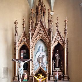 Zdjęcie nr 1: Ołtarz przyścienny, architektoniczny, trójosiowy, jednokondygnacyjny ze zwieńczeniem. Mensa skrzyniowa, prostopadłościenna. Antepedium z trzema prostokątnymi płycinami wypełnionymi ornamentem roślinnym. Predella wysoka, dekorowana fryzem arkadkowym, ostrołukowym. Retabulum jest podzielone czterema smukłymi kolumnami zamkniętymi sterczynami z żabkami i kwiatonem. Pole środkowe retabulum wyższe i szersze w formie prostokątnej wnęki zamkniętej łukiem ostrym, w osiach bocznych ostrołukowe nisze zakończone wimpergą z rozetą. Ołtarz ma zwieńczenie w formie ażurowej wieży z ostrołukowymi arkadkami zwieńczonej żabkami i kwiatonem, po bokach sterczyny z kwiatonem. W polu środkowym retabulum obraz z przedstawieniem św. Teresy z Lisieux ujęty w ościeża dekorowane ornamentem w formie różanych gałązek i kwiatów. W niszach bocznych pełnoplastyczne, polichromowane figury św. Stanisława Kostki (?) i św. Franciszka ustawione na dekoracyjnych cokołach ze sterczynami na narożach i płyciną z ornamentem roślinnym od frontu. Tabernakulum w formie prostopadłościennej szafki z drzwiczkami ozdobionymi płyciną, w której płaskorzeźbiony kielich z hostią na obłokach. Po bokach drzwiczek płyciny z ornamentem uformowanym z różanych gałązek. Tron wystawienia w formie baldachimu z dwuspadowym dachem wspartym na dwóch kolumnach, zwieńczonych sterczynami z żabkami i kwiatonami.  