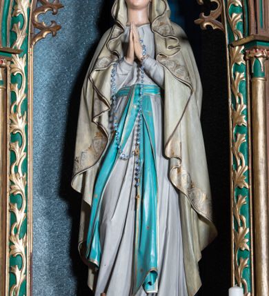 Zdjęcie nr 1: Rzeźba pełna na cokole przedstawiająca stojącą frontalnie Matkę Boską z lekko ugiętą prawą nogą i rękami złożonymi do modlitwy; przez dłonie ma przełożony różaniec. Maria ma lekko uniesioną głowę, oczy skierowane w górę, nos prosty, usta wąskie. Jest ubrana w białą suknię przepasaną niebieskim pasem ze złotą lamówką, którego długie końce opadają w dół. Ma również narzucony na głowę oraz ramiona, opadający miękkimi fałdami płaszcz dekorowany na brzegach pasem złotego ornamentu. Spod sukni wystają bose stopy, na każdej z nich kwiat róży. 
Polichromia w partiach ciała naturalistyczna. 