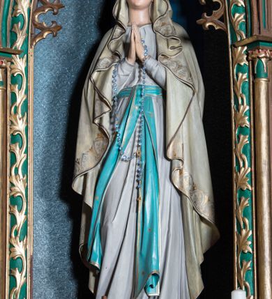 Zdjęcie nr 1: Rzeźba pełna na cokole przedstawiająca stojącą frontalnie Matkę Boską z lekko ugiętą prawą nogą i rękami złożonymi do modlitwy; przez dłonie ma przełożony różaniec. Maria ma lekko uniesioną głowę, oczy skierowane w górę, nos prosty, usta wąskie. Jest ubrana w białą suknię przepasaną niebieskim pasem ze złotą lamówką, którego długie końce opadają w dół. Ma również narzucony na głowę oraz ramiona, opadający miękkimi fałdami płaszcz dekorowany na brzegach pasem złotego ornamentu. Spod sukni wystają bose stopy, na każdej z nich kwiat róży. 
Polichromia w partiach ciała naturalistyczna. 