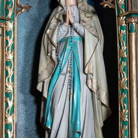 Zdjęcie nr 1: Rzeźba pełna na cokole przedstawiająca stojącą frontalnie Matkę Boską z lekko ugiętą prawą nogą i rękami złożonymi do modlitwy; przez dłonie ma przełożony różaniec. Maria ma lekko uniesioną głowę, oczy skierowane w górę, nos prosty, usta wąskie. Jest ubrana w białą suknię przepasaną niebieskim pasem ze złotą lamówką, którego długie końce opadają w dół. Ma również narzucony na głowę oraz ramiona, opadający miękkimi fałdami płaszcz dekorowany na brzegach pasem złotego ornamentu. Spod sukni wystają bose stopy, na każdej z nich kwiat róży. 
Polichromia w partiach ciała naturalistyczna. 