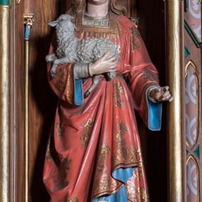 Zdjęcie nr 1: Rzeźba pełna na cokole przedstawiająca stojącą frontalnie św. Agnieszkę ukazaną w lekkim kontrapoście. Głowa delikatnie uniesiona, oczy skierowane w górę, nos prosty, usta wąskie. Brązowe, falowane włosy spływają na ramiona i plecy. Na głowie ma złotą koronę otwartą. Lewa ręka lekko zgięta i wysunięta do przodu w delikatnym geście, prawa przyciska do piersi baranka spoczywającego na przedramieniu. Święta Agnieszka jest ubrana w spodnią, białą suknię lamowaną pod szyją złotem oraz wierzchnią czerwoną ze złotymi motywami kwiatowymi i niebieskim podbiciem, przepasaną i podciągniętą z lewej strony. Spod szat wystają noski brązowych butów. Polichromia w partiach ciała naturalistyczna.