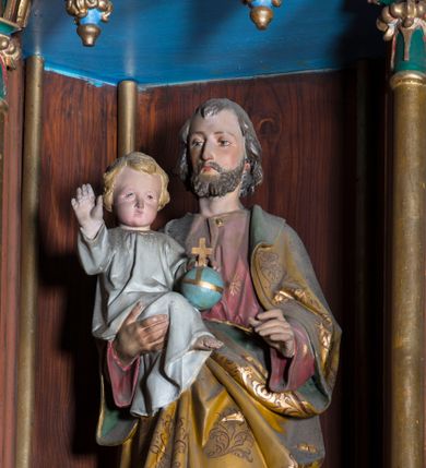 Zdjęcie nr 1: Rzeźba pełna na cokole przedstawiająca stojącego frontalnie św. Józefa z Dzieciątkiem Jezus na ręku. Święty został ujęty w lekkim kontrapoście. Ma głowę ustawioną prosto, oczy skierowane przed siebie, nos prosty, usta wąskie; ciemny zarost oraz włosy zakrywające uszy i opadające na kark. Lewą rękę ma lekko zgiętą i wysuniętą do przodu,  prawą podtrzymuje siedzące Dzieciątko Jezus. Święty Józef jest ubrany w czerwoną suknię, na którą ma narzucony żółty płaszcz dekorowany motywami roślinnymi, z zielonym podbiciem. Dzieciątko prawą rękę unosi do góry, lewą przytrzymuje jabłko królewskie. Ma owalną twarz z wysokim czołem, małymi oczami i ustami oraz wąskim nosem. Ma krótkie, jasne włosy z przedziałkiem, jest ubrane w długą, białą sukienkę. Polichromia w partiach ciała naturalistyczna.