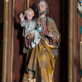 Zdjęcie nr 1: Rzeźba pełna na cokole przedstawiająca stojącego frontalnie św. Józefa z Dzieciątkiem Jezus na ręku. Święty został ujęty w lekkim kontrapoście. Ma głowę ustawioną prosto, oczy skierowane przed siebie, nos prosty, usta wąskie; ciemny zarost oraz włosy zakrywające uszy i opadające na kark. Lewą rękę ma lekko zgiętą i wysuniętą do przodu,  prawą podtrzymuje siedzące Dzieciątko Jezus. Święty Józef jest ubrany w czerwoną suknię, na którą ma narzucony żółty płaszcz dekorowany motywami roślinnymi, z zielonym podbiciem. Dzieciątko prawą rękę unosi do góry, lewą przytrzymuje jabłko królewskie. Ma owalną twarz z wysokim czołem, małymi oczami i ustami oraz wąskim nosem. Ma krótkie, jasne włosy z przedziałkiem, jest ubrane w długą, białą sukienkę. Polichromia w partiach ciała naturalistyczna.